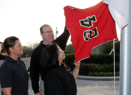 Jaguars Softball Coach Keri Lambeth, left, watches on Monday morning as Verizon Wireless Manager Casey Houseworth and customer service agent Brunilda Ortiz inspect the Jaguar Jersey before raising it up the UHV flagpole.
